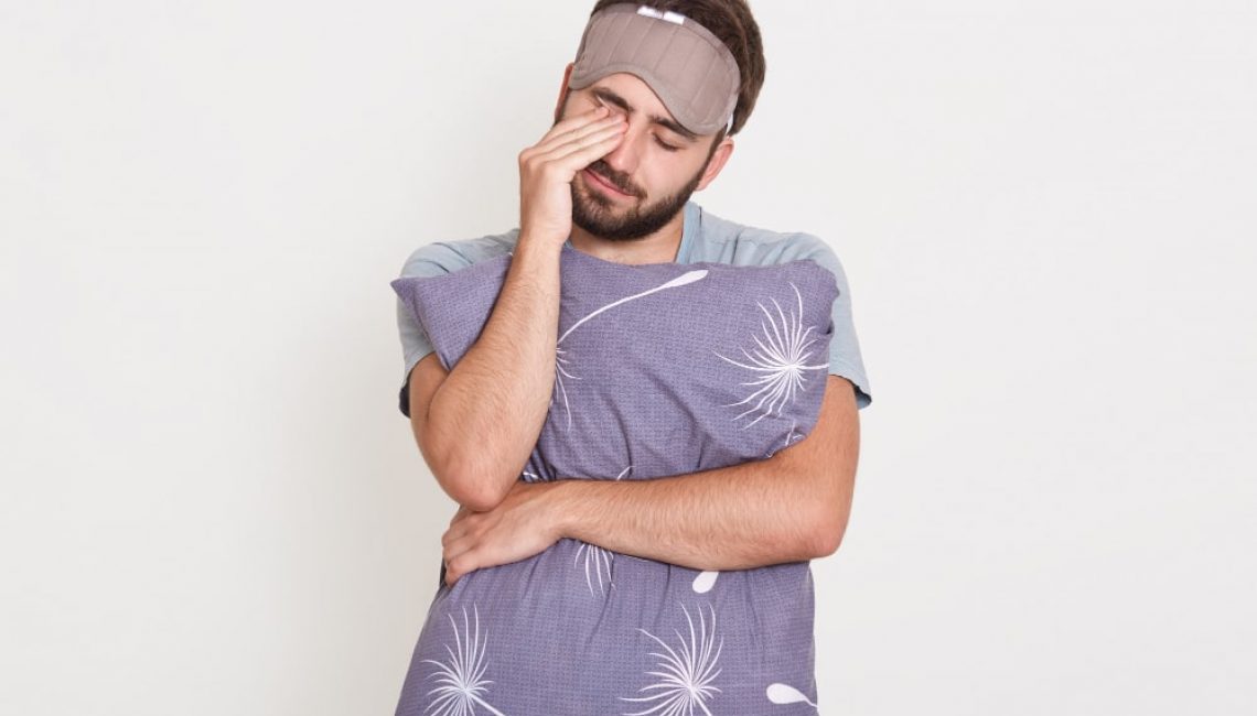 sleepy-bearded-male-posing-with-closed-eyes-isolated-over-white-wall-rubbing-his-eyes-with-hand-embracing-pillow-min